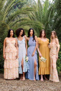 four women in dresses standing next to each other with palm trees and bushes behind them