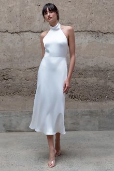 a woman standing in front of a stone wall wearing a white dress with high neck