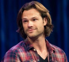 a man with long hair wearing a red and black flannel shirt is standing in front of a blue curtain