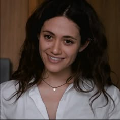 a woman with long dark hair wearing a white shirt and smiling at the camera while standing in front of a wood paneled wall