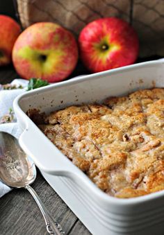 a casserole dish with apples in the background and two spoons next to it