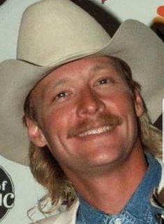 a man with long hair wearing a white cowboy hat and blue shirt smiling at the camera