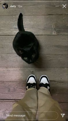 a black cat sitting on top of a wooden floor next to someone's feet