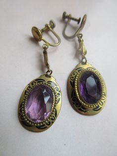 Vintage Deco long drop screw back earrings. These estate earrings are crafted of a brassy metal and set with an amethyst colored faceted glass. The stones are bezel set with black tracery embellishment frames. The stones are slightly different. These open back drops are oval in form and measure seven eighths of an inch by five eighths of an inch. The drops have a total length of 1.5''.  Truly a great vintage pair that will compliment your Art Deco Era jewelry collection.  age appropriate wear Purple Vintage Jewelry, Purple Brass Jewelry With Matching Earrings, Antique Purple Metal Jewelry, Vintage Purple Teardrop Jewelry, Formal Purple Clip-on Jewelry, Faceted Briolette Amethyst Earrings, Purple Brass Drop Earrings, Faceted Amethyst Dangle Jewelry, Vintage Purple Teardrop Earrings