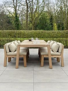 a wooden table with four chairs around it in front of some bushes and trees on the other side