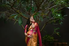 a woman in an orange and pink sari standing next to a tree with green leaves