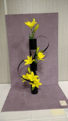 a black vase with yellow flowers in it on a purple tableclothed surface next to a white wall