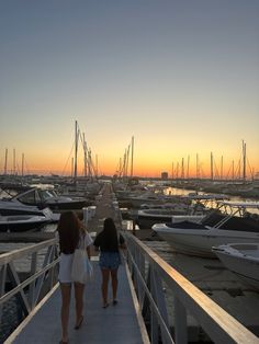 two women are walking down a dock towards boats at the marina as the sun sets