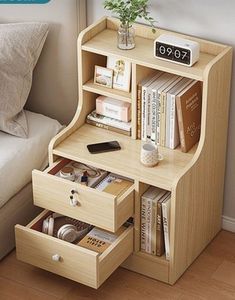 a small wooden desk with drawers and books on it in front of a bed next to a window