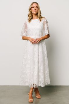 a woman wearing a white dress with sheer sleeves and floral embroidery on the shoulders, standing in