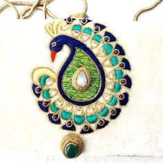 an embroidered peacock ornament hanging on a white cloth with gold trimmings