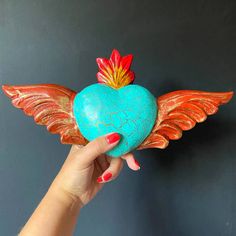 a hand holding a heart shaped object with wings on the top, and an orange flower in the middle