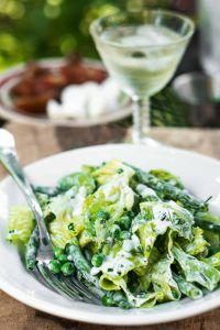 a white plate topped with pasta covered in peas and sauce next to a glass of wine