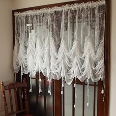 a wooden chair sitting in front of a window covered in white lace curtaines and tassels