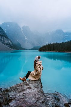 a woman sitting on top of a rock next to a lake