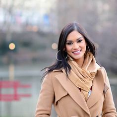 a woman in a tan coat is smiling and holding her hand on her hip as she walks down the street