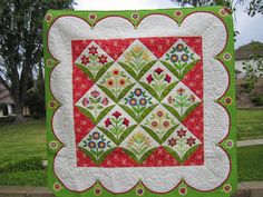 a green and red quilt with flowers on it hanging from a brick wall in front of some trees