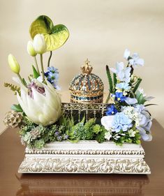 an arrangement of flowers in a white vase on a table with a gold crown and jeweled box