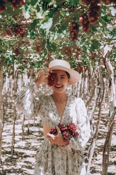 a woman in a white dress and straw hat is holding grapes up to her face