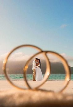 a man and woman standing on top of a beach next to the ocean with rings in front of them