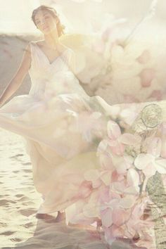 a woman in a white dress is standing on the beach with flowers and clouds behind her