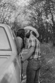 a man and woman kissing by the back of a car in front of some trees