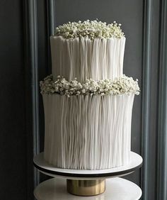 a three tiered cake with white flowers on top is sitting on a stand in front of a gray wall