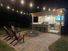 an outdoor bar and grill with lights strung over it at night, next to two adiron chairs