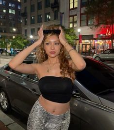 a woman standing in front of a car with her hands on her head and looking at the camera