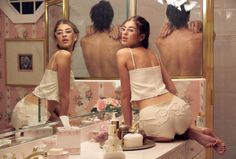 two women sitting on a counter in front of a mirror looking at themselves in the bathroom