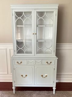 a white china cabinet with glass doors and gold knobs on the bottom, against a beige wall