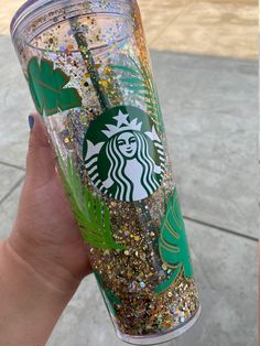 a starbucks cup with gold glitters and green leaves on the lid is being held up