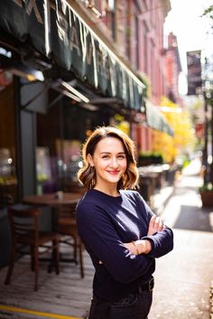 a woman standing on the sidewalk in front of a restaurant with her arms crossed and smiling