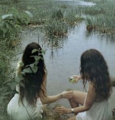 two young women sitting on the ground in front of a body of water and plants