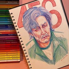 colored pencils next to a drawing of a man's face on a notebook