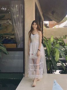 a woman in a white dress standing next to a wall with plants behind her and a blue pillow on the floor