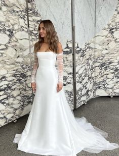 a woman standing in front of a marble wall wearing a white wedding dress with long sleeves