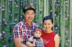 a man and woman holding a baby in front of a bamboo fence with purple flowers