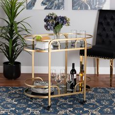 a gold bar cart with wine glasses and plates on it next to a potted plant