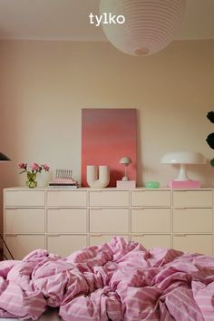 a bed with pink sheets and pillows in a bedroom next to a dresser, lamp and painting on the wall