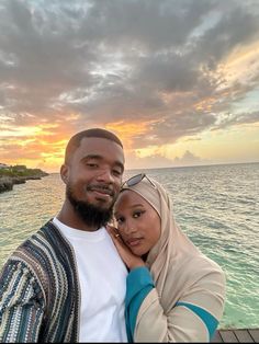 a man and woman standing next to each other in front of the ocean at sunset