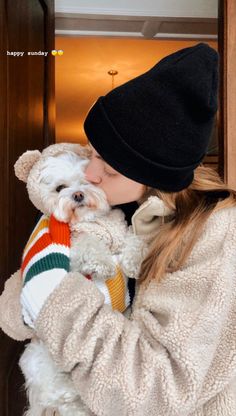 a woman holding a small white dog wearing a hat and sweater in front of a door