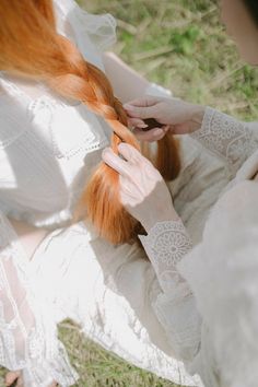 a woman with long red hair is sitting on the grass and braiding her hair