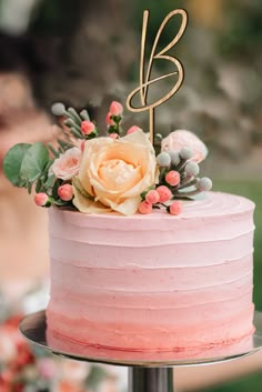 a pink frosted cake with flowers on top and greenery around the edges is sitting on a metal stand