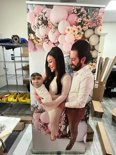 a man and woman holding a baby standing in front of a large poster with balloons on it