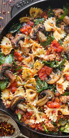 a skillet filled with pasta and vegetables on top of a wooden table next to spices