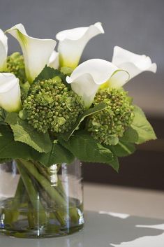 there is a vase with white flowers and green leaves in it on top of a table