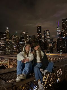 two young women sitting on a bridge in front of the city skyline at night time