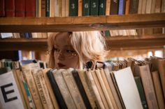 a blonde haired woman with glasses looking at books in a book store, from behind the shelves