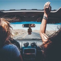 two women are driving down the road with their hands in the rear view mirror and one is holding up her hand